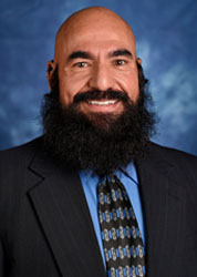 a bald man with a medium length black beard wearing a black suit jacket with a blue shirt and black and blue tie standing in front of a blue background.