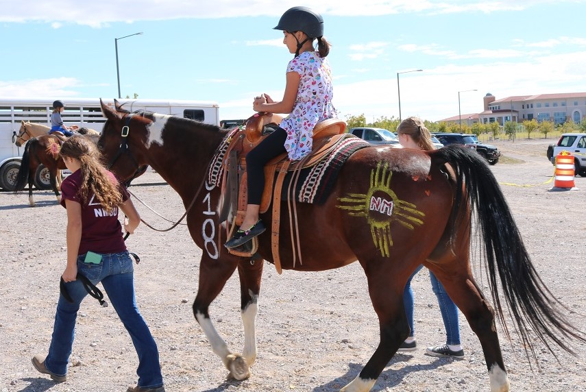 Dos mujeres en edad universitaria guían un caballo marrón montado por una niña con casco.