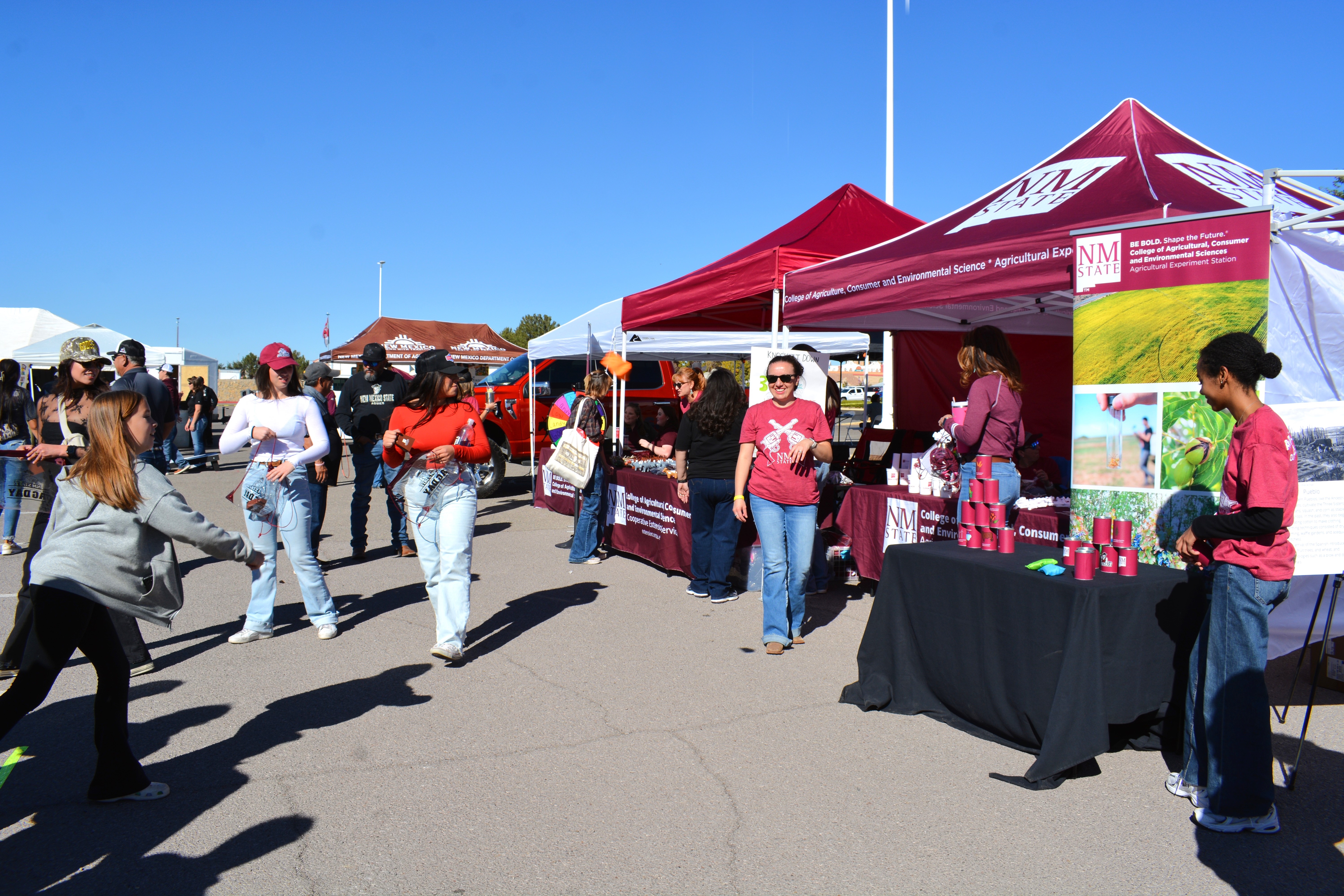 People engaging with booths at a community event, with tents displaying information and products from New Mexico State University.