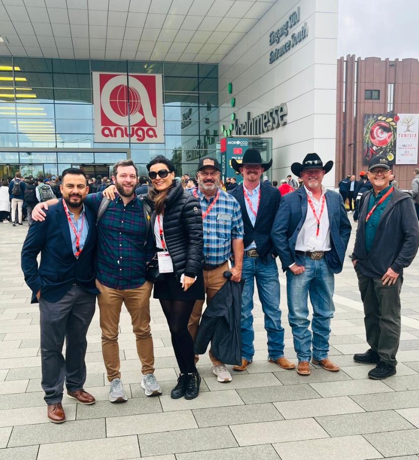 A group of eight people, including New Mexico producers and trade partners, pose outside the ANUGA food trade show entrance in Cologne, Germany.