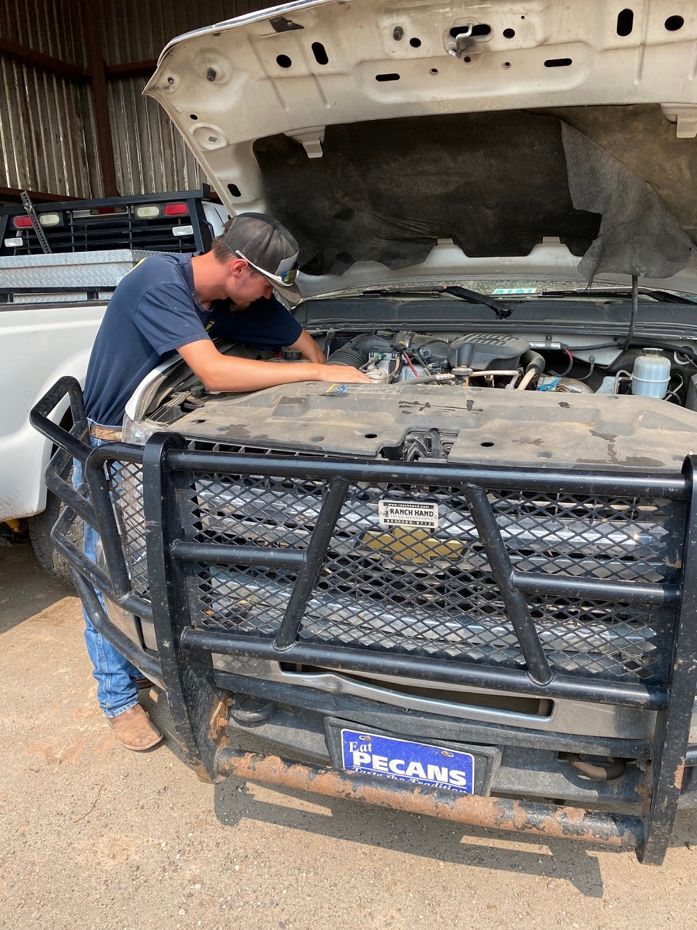 A young man with a baseball cap stands over a truck with the hood propped open, working on the engine.