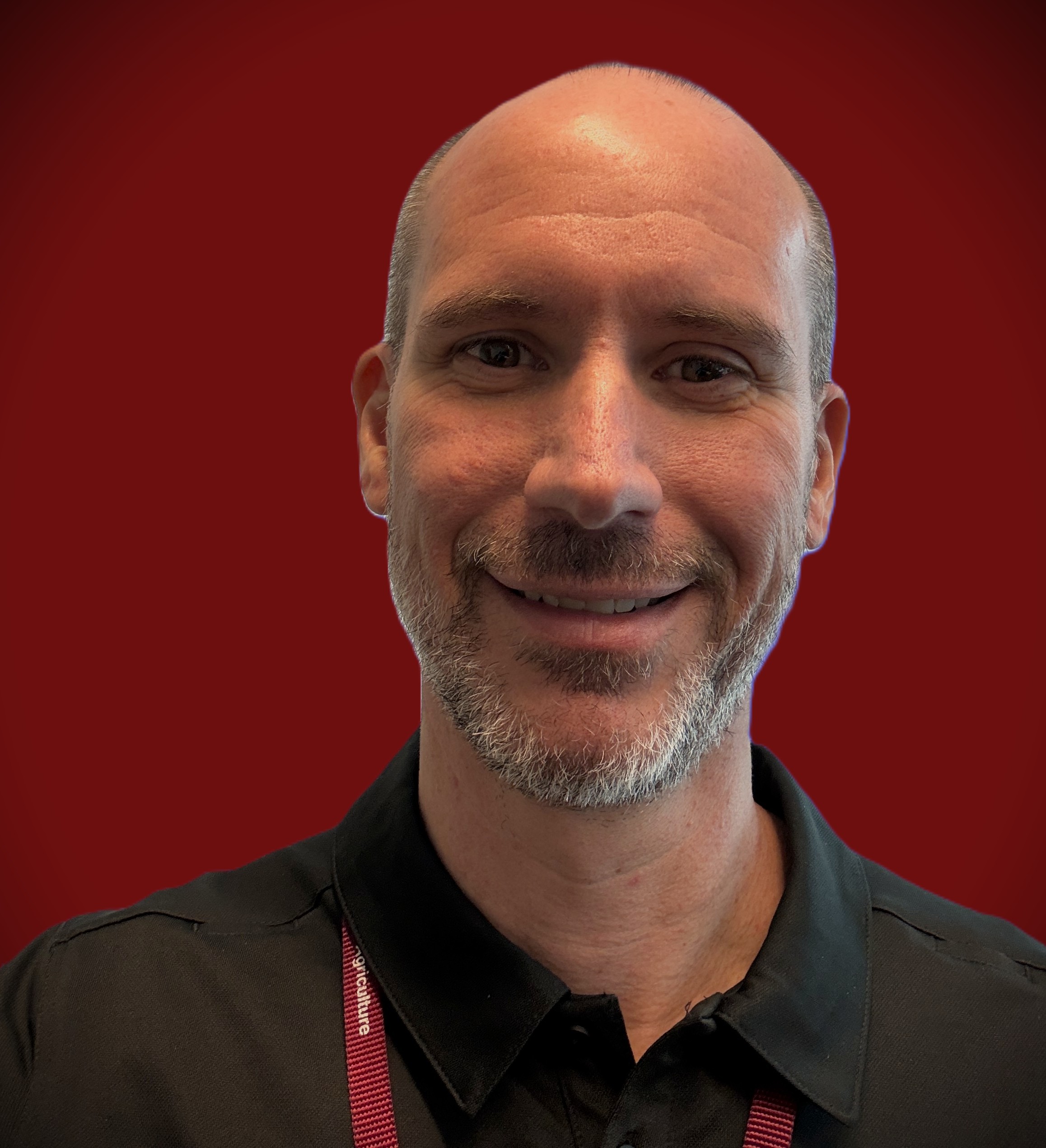 Headshot of a smiling man with closely cropped hair and a short beard, wearing a black collared shirt against a red background.