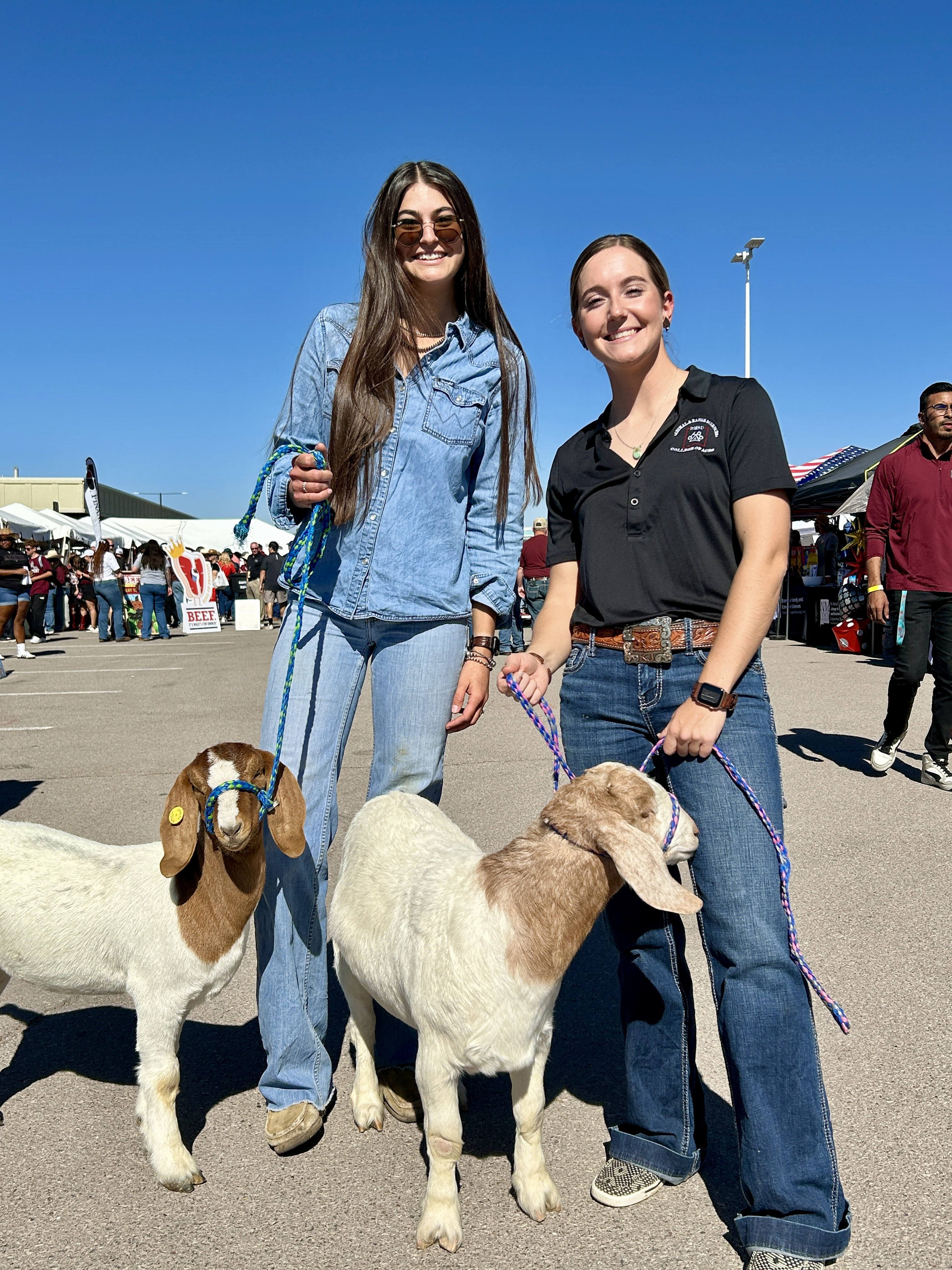 Two women pose with two sheep at an outdoor agricultural event.