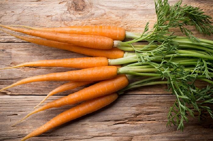 Several orange carrots with long green leafy stems lay horizontally on a brown wooden table.