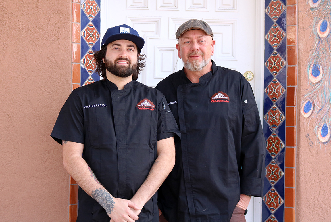 A man on the left with dark brown hair and beard wears a black short-sleeve chef coat and blue baseball cap. A man on the right with a white beard and gray hat wears a long-sleeve black chef coat. The backdrop is a white door framed by blue and orange decorative tile.