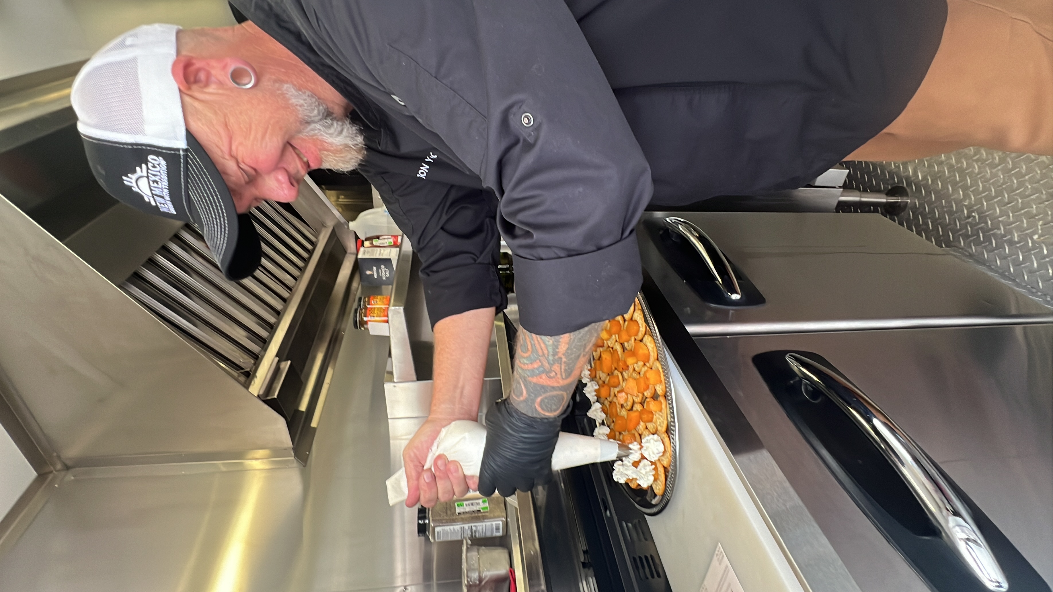 A male chef in a black coat putting toppings on food samples. 