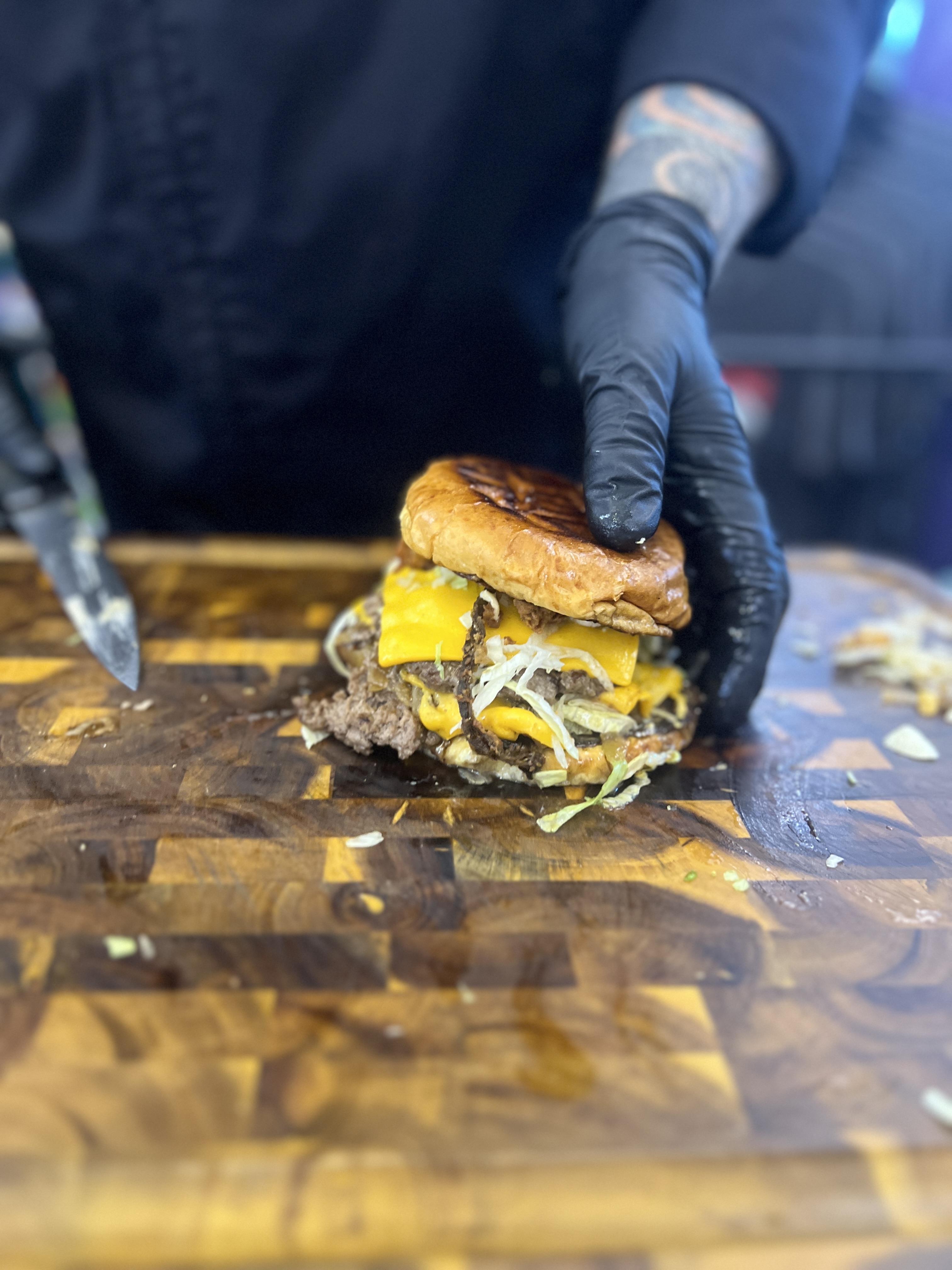 A green chile cheeseburger sits on a checkered cutting board. A chef in a black coat wearing black gloves holds the burger on one side. The chef can be seen holding a knife in the other hand.