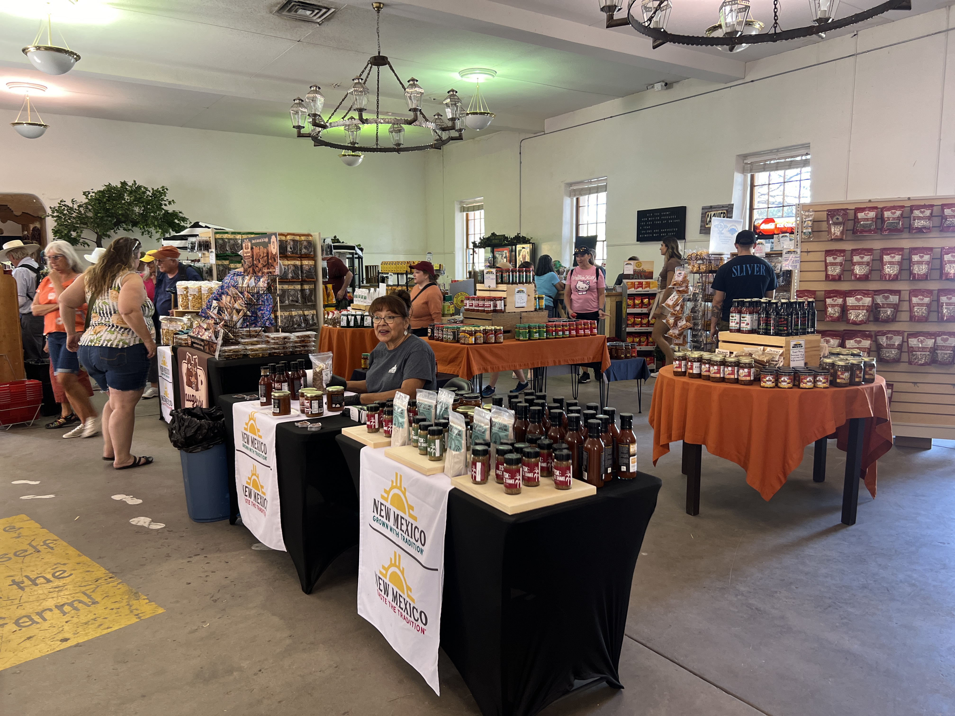 Fairgoers browse tables filled with New Mexico-made food products inside the Country Store at the New Mexico State Fair. A vendor sits behind a table displaying items with the “New Mexico–Taste the Tradition” banner.