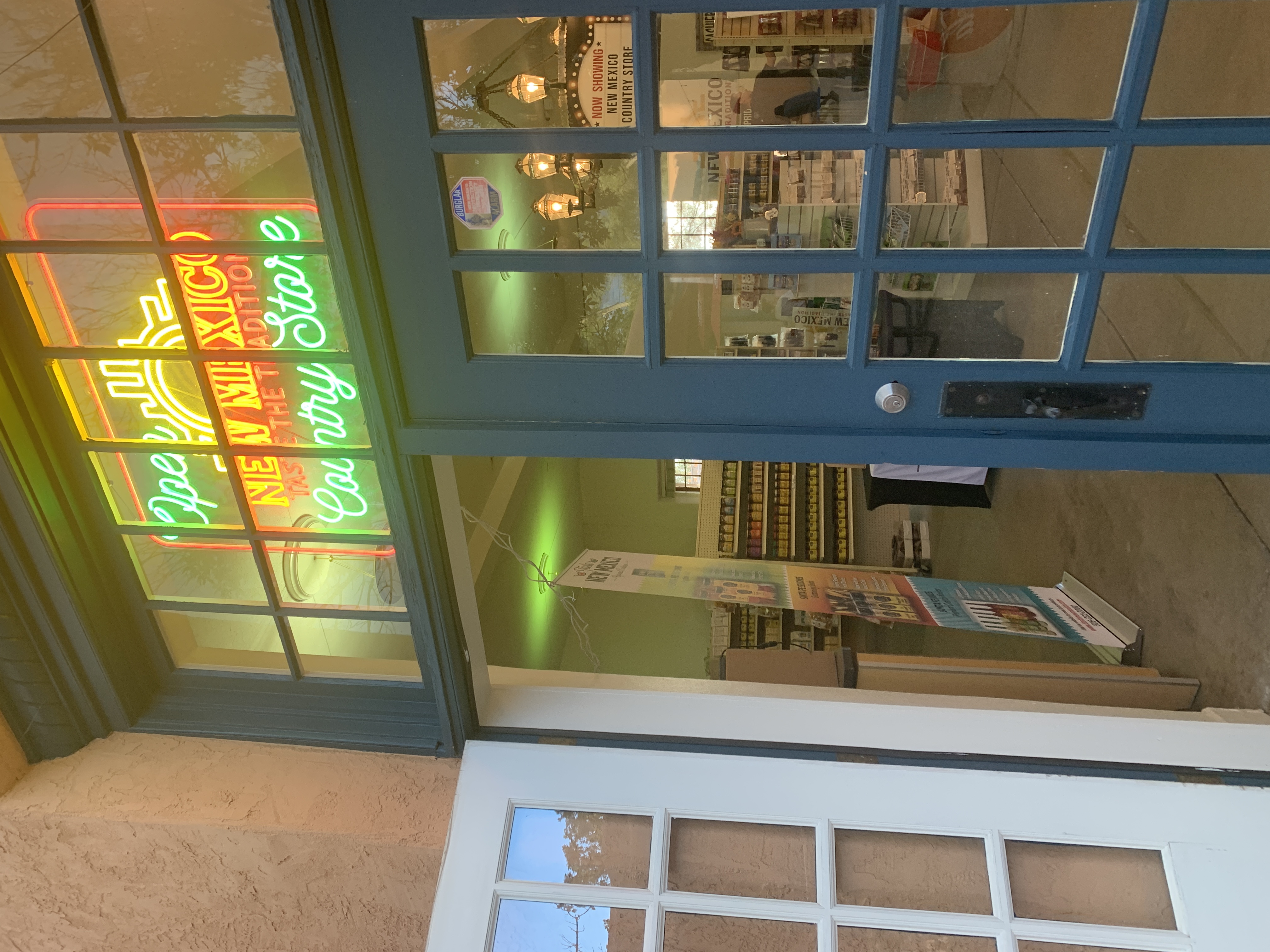 Store entrance with a blue-framed glass door and an “Open New Mexico Taste the Tradition Country Store” neon sign glowing above the windows, showing shelves of products inside.