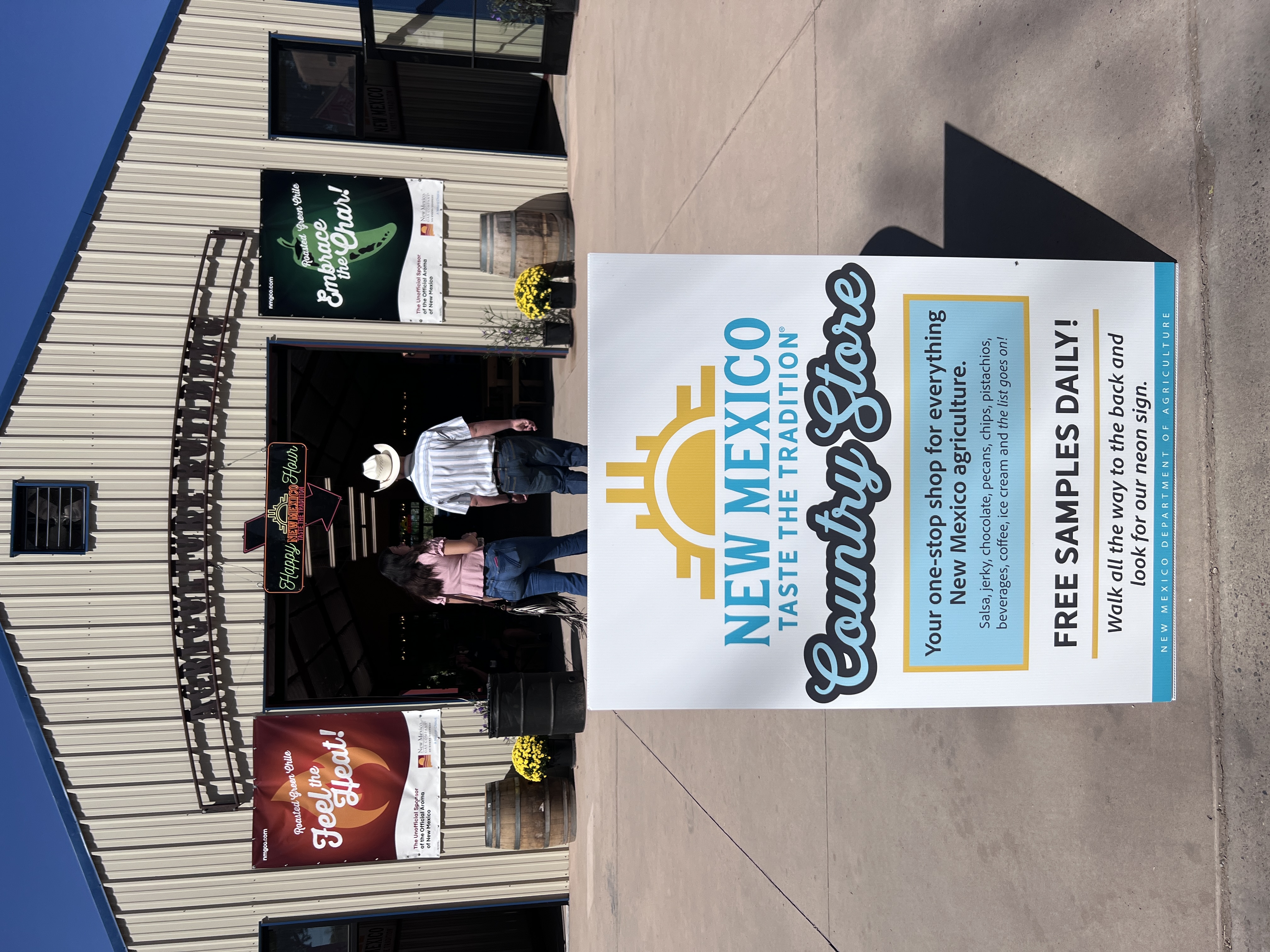 A large outdoor sign reads “New Mexico Taste the Tradition Country Store” with a list of local products and free samples. Fairgoers walk toward the entrance of the Agriculture Building under signs promoting New Mexico chile.