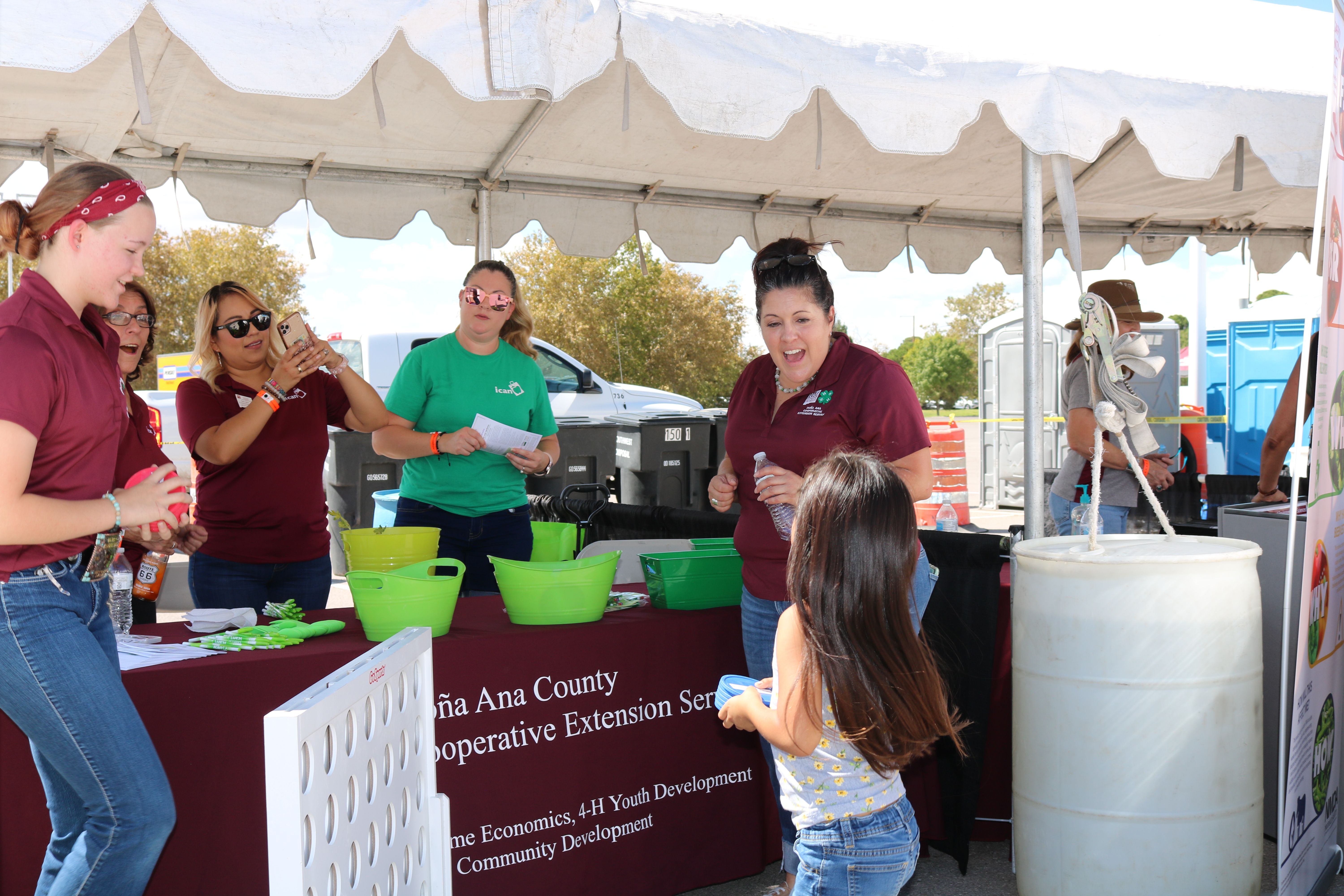 Five women cheer on a young girl playing a game of Connect Four. A table with a crimson tablecloth and green buckets is in the background. 