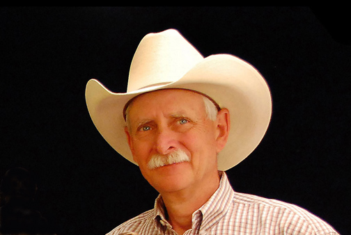 A man with a white cowboy hat and a striped shirt hiding a smile through his white mustache with a black background. 