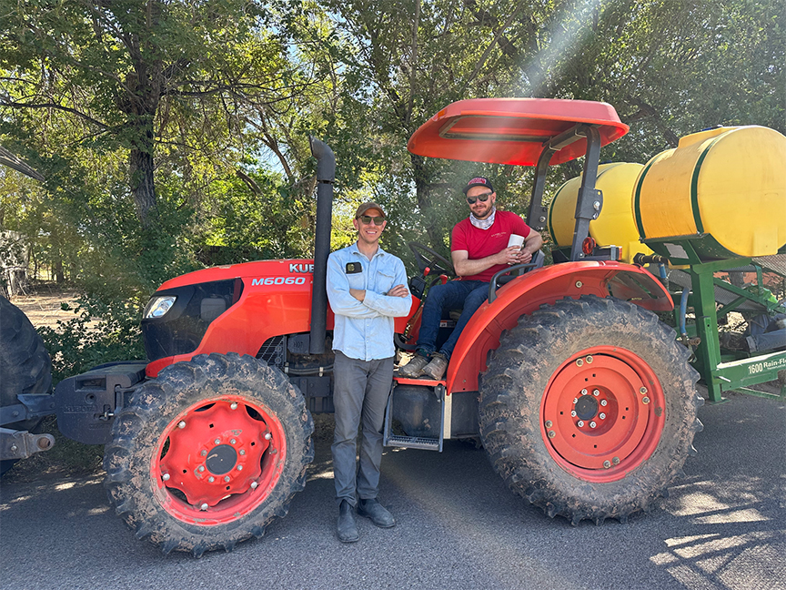 Two men standing with a red Kubota tractor and sprayer on a sunny day.