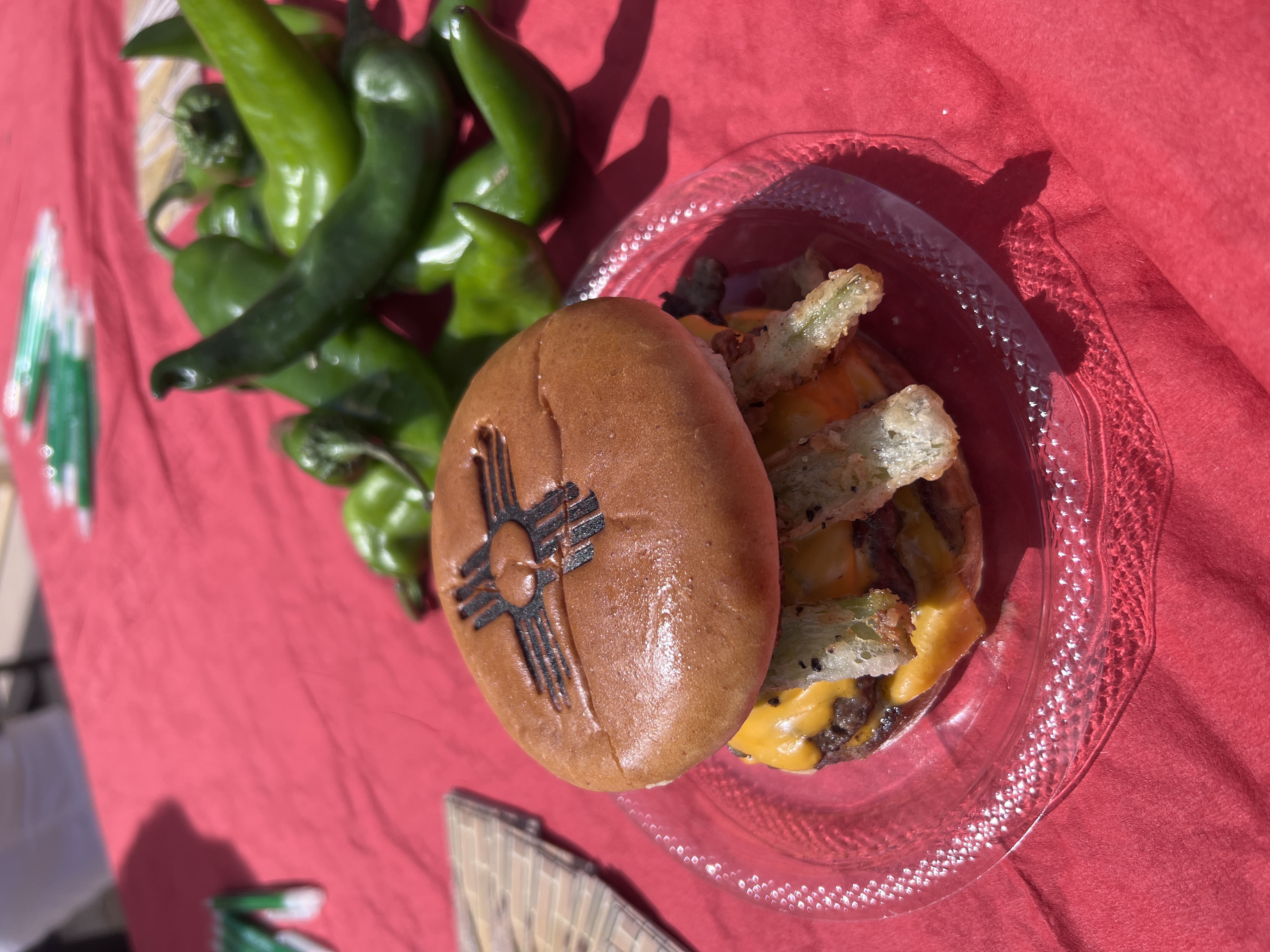 A cheeseburger with green chile and the New Mexico Zia logo branded on top of it, next to a handful of raw green chiles. 