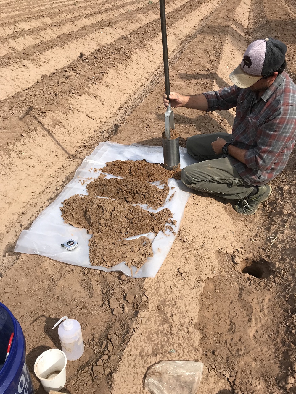 Un hombre se arrodilla en un campo de cultivo desnudo mientras recolecta muestras de suelo con un gran instrumento de plata.