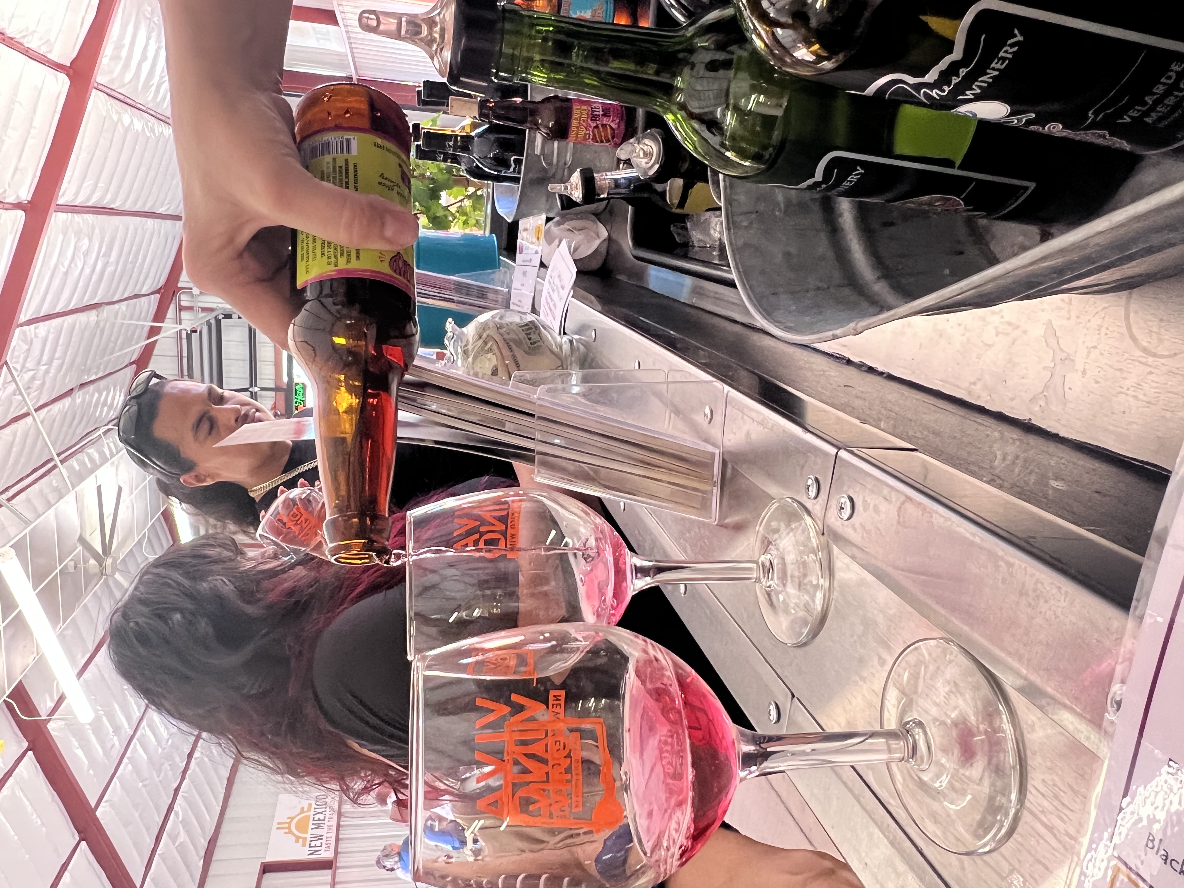 A vendor pours rosé wine into glasses at the Happy Hours wine bar during the New Mexico State Fair. Guests watch from across the counter, surrounded by local wine bottles and signage for “Viva Vino.”