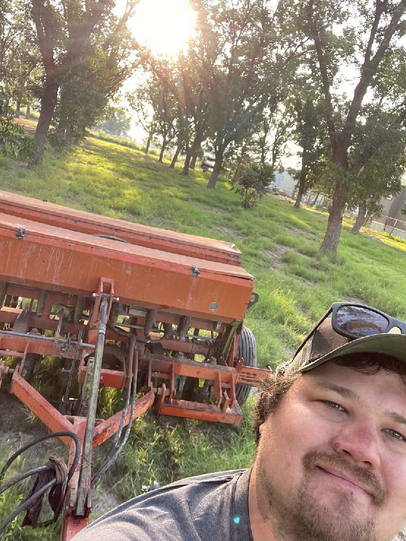 A man wearing a baseball cap is standing in front of red farm equipment, known as a seed drill. Grass and a pecan orchard are behind him with the sun shining through the tree tops.
