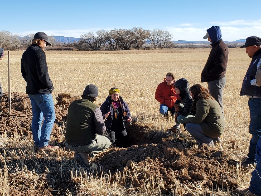 Una mujer se para en un gran hoyo excavado en un campo de cultivo inactivo, mientras siete personas la rodean mientras habla.