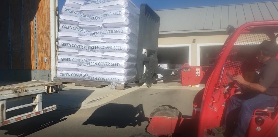 A man driving a red forklift unloads a pallet with several large white bags labeled “Green Cover Seed” from a box truck. 