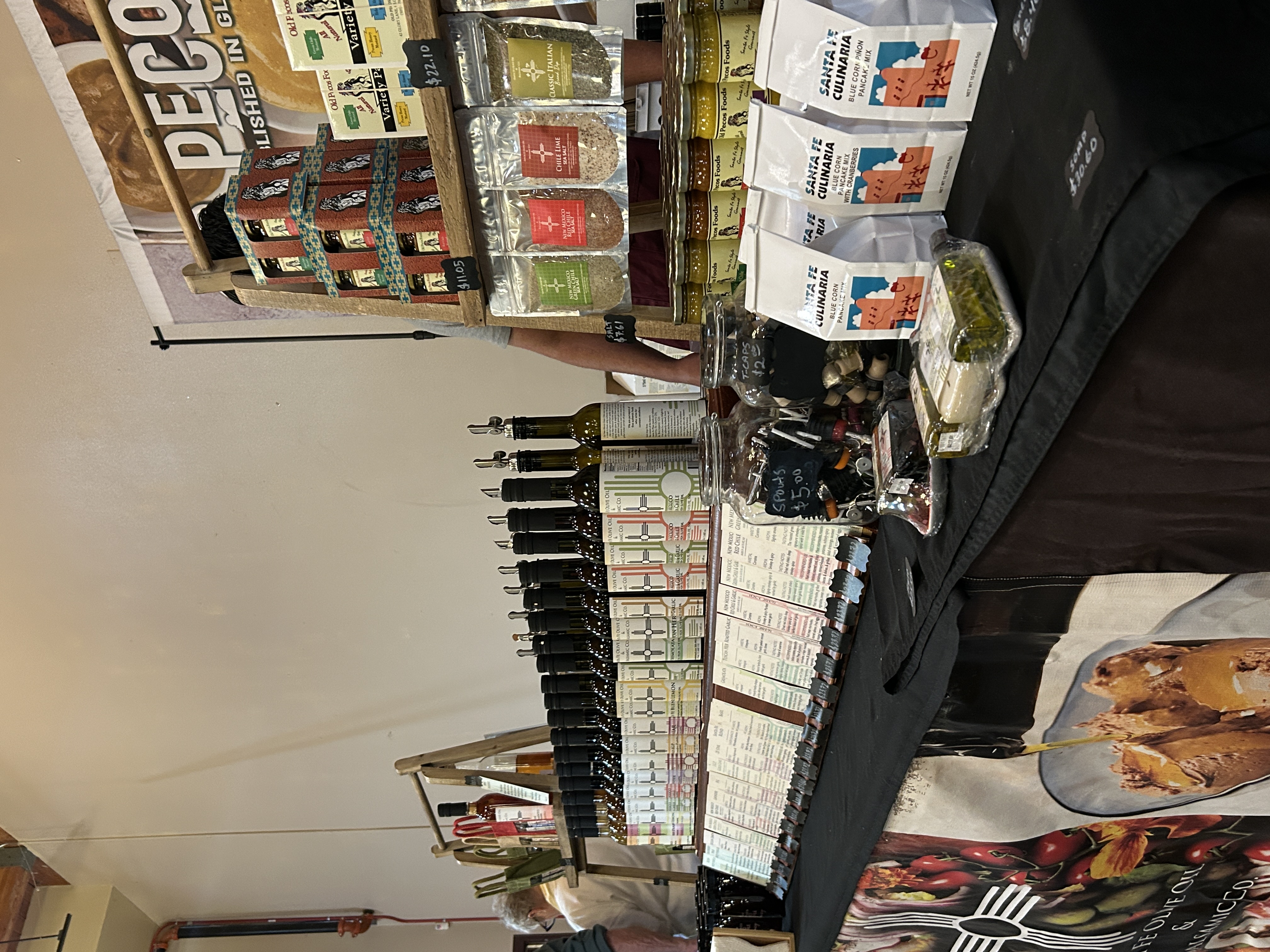 The Pecos Foods vendor booth at the HomeGrown event features rows of bottled oils and sauces, jars of preserves, seasoning blends, and packaged baking mixes displayed on a black tablecloth beneath a sign promoting the brand.