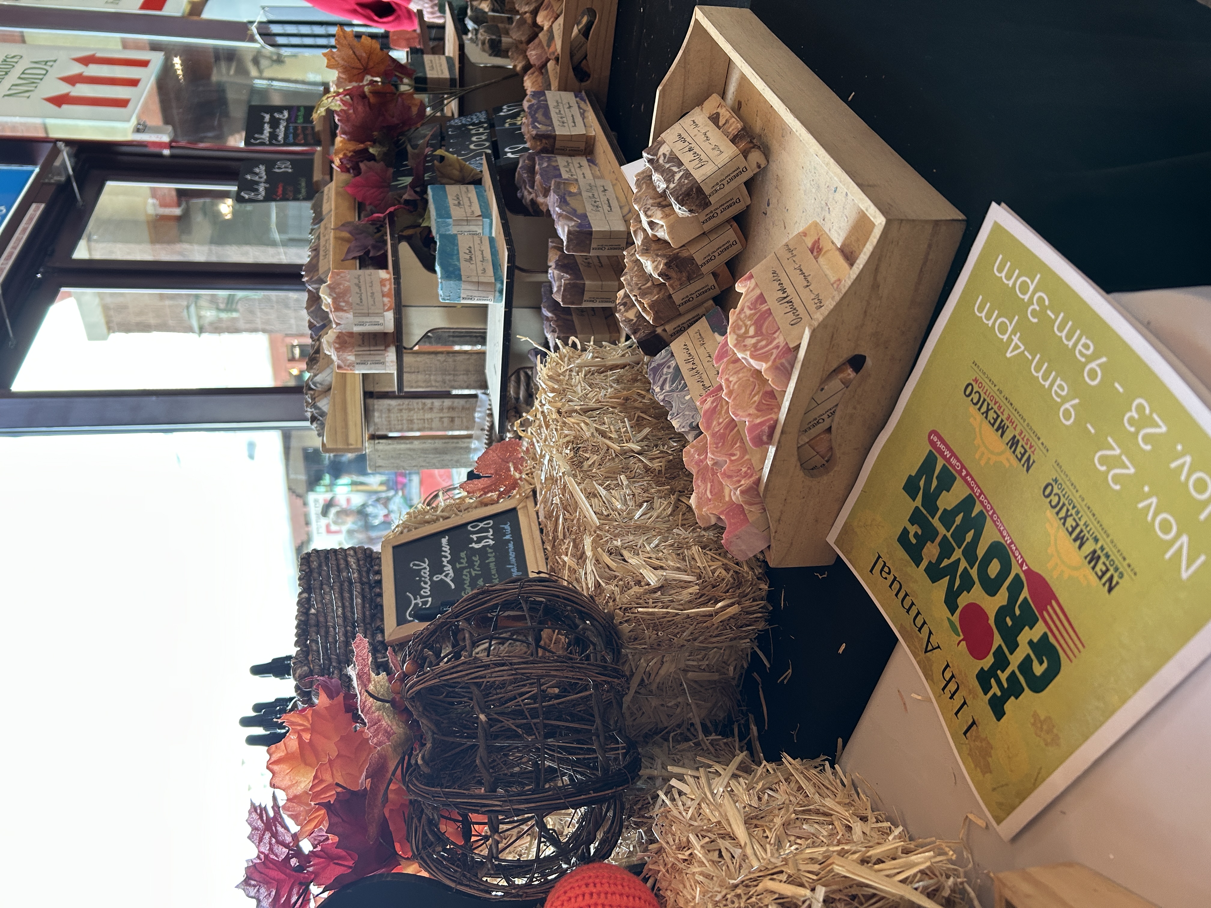 A display of handmade soaps arranged on hay bales and wooden trays, decorated with autumn leaves and rustic pumpkins. A flyer for the 11th annual HomeGrown event, scheduled for Nov. 22–23, sits in the foreground