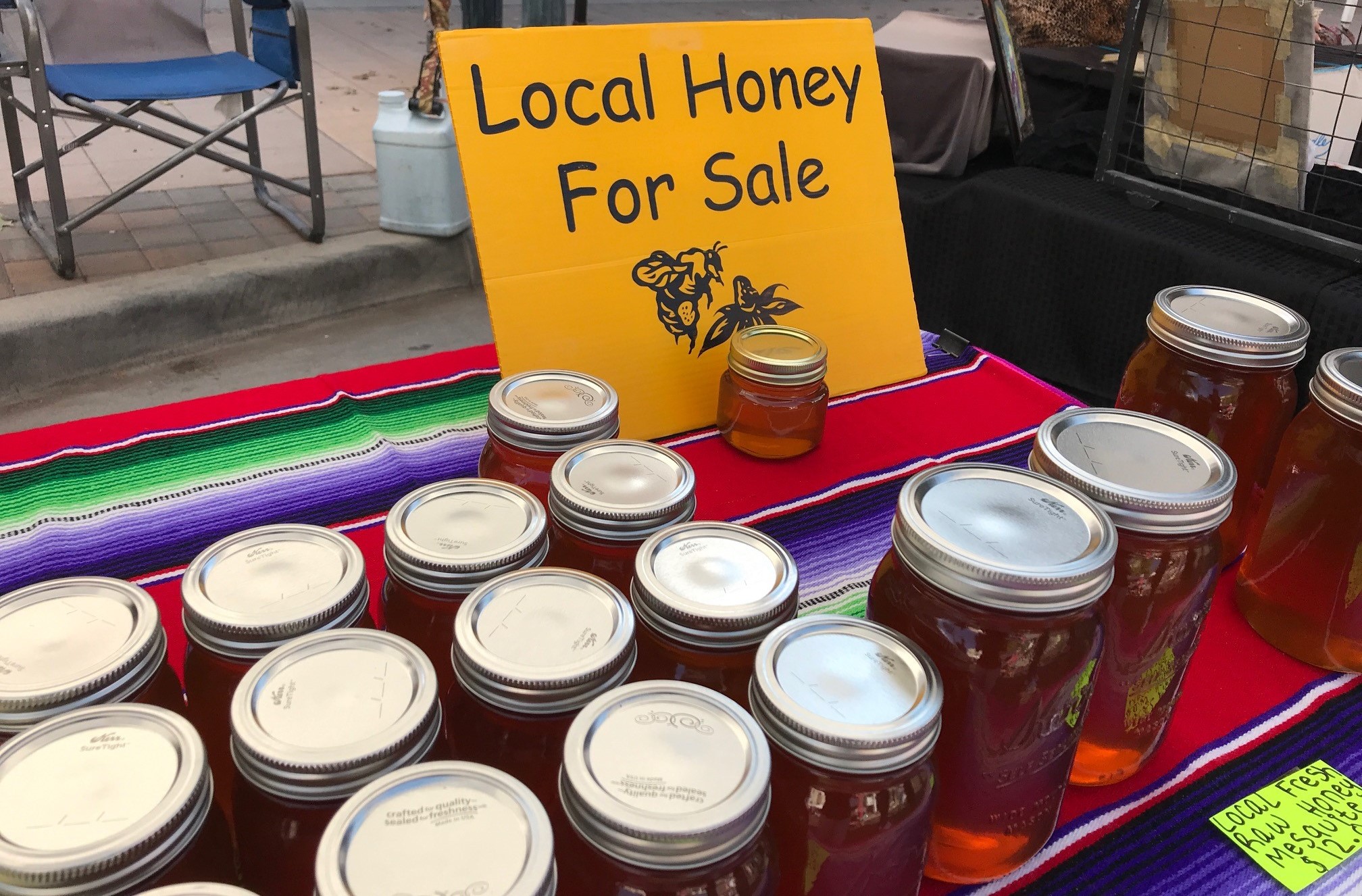 Several jars of honey on a table with colorful table cloth, with yellow sign with black lettering reading “Local Honey For Sale” behind the jars