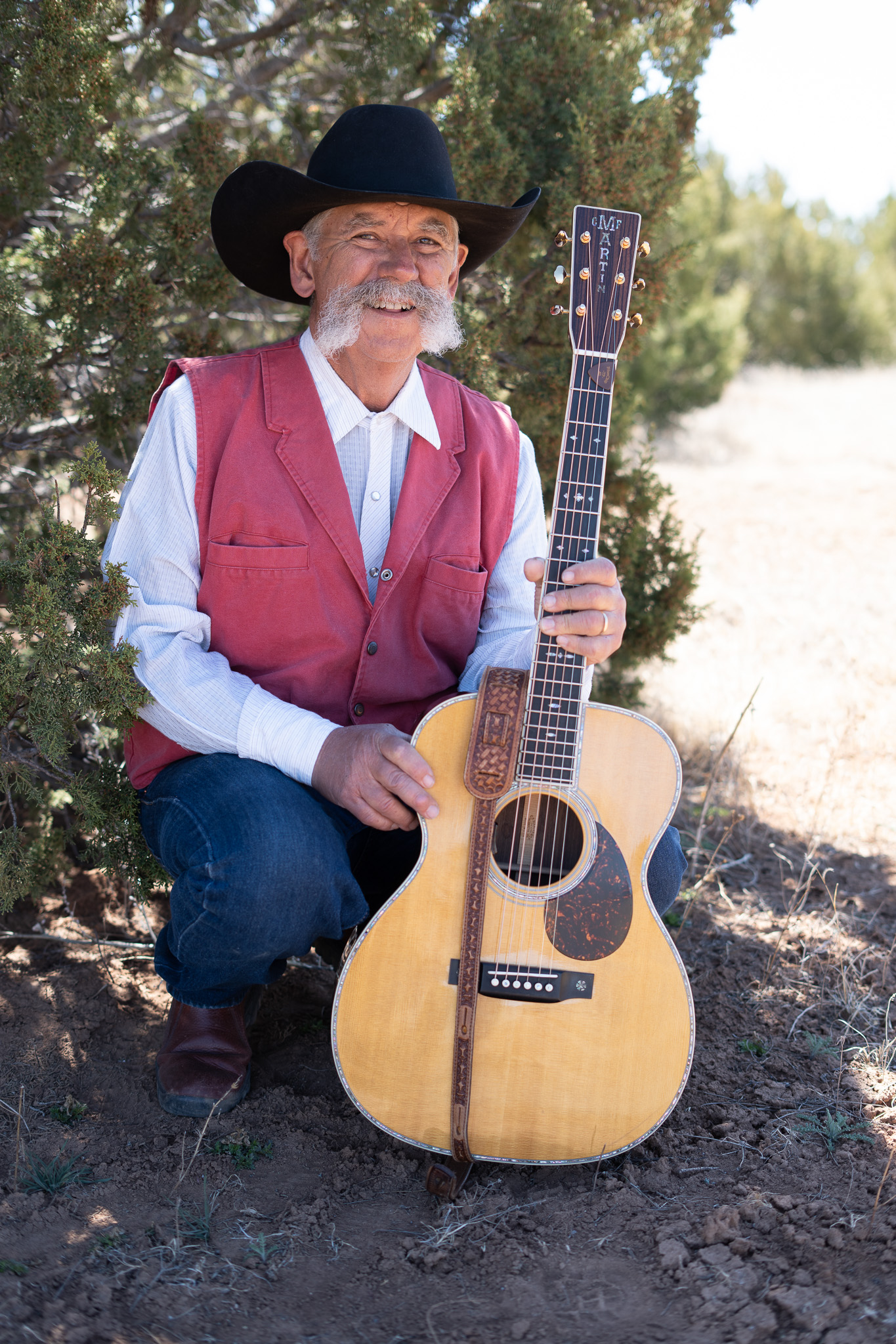 A man in a cowboy hat and red vest posing with an acoustic guitar outside.