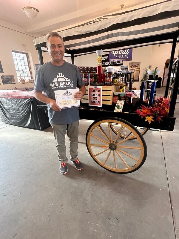 A man holding a sign that reads “I am grateful for New Mexico agriculture” in front of a display wagon of salsa products inside the country store.