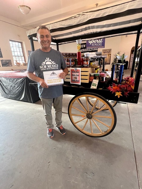 Un hombre con un letrero que dice “Estoy agradecido por la agricultura de Nuevo México” frente a un carrito de exhibición de productos de salsa dentro de la tienda.