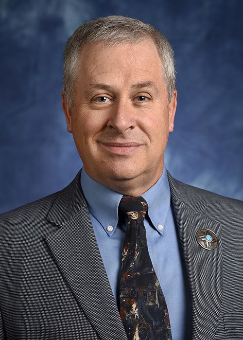 a man with short grey hair standing in front of a blue background wearing a blue shirt, grey suit and dark blue tie. 
