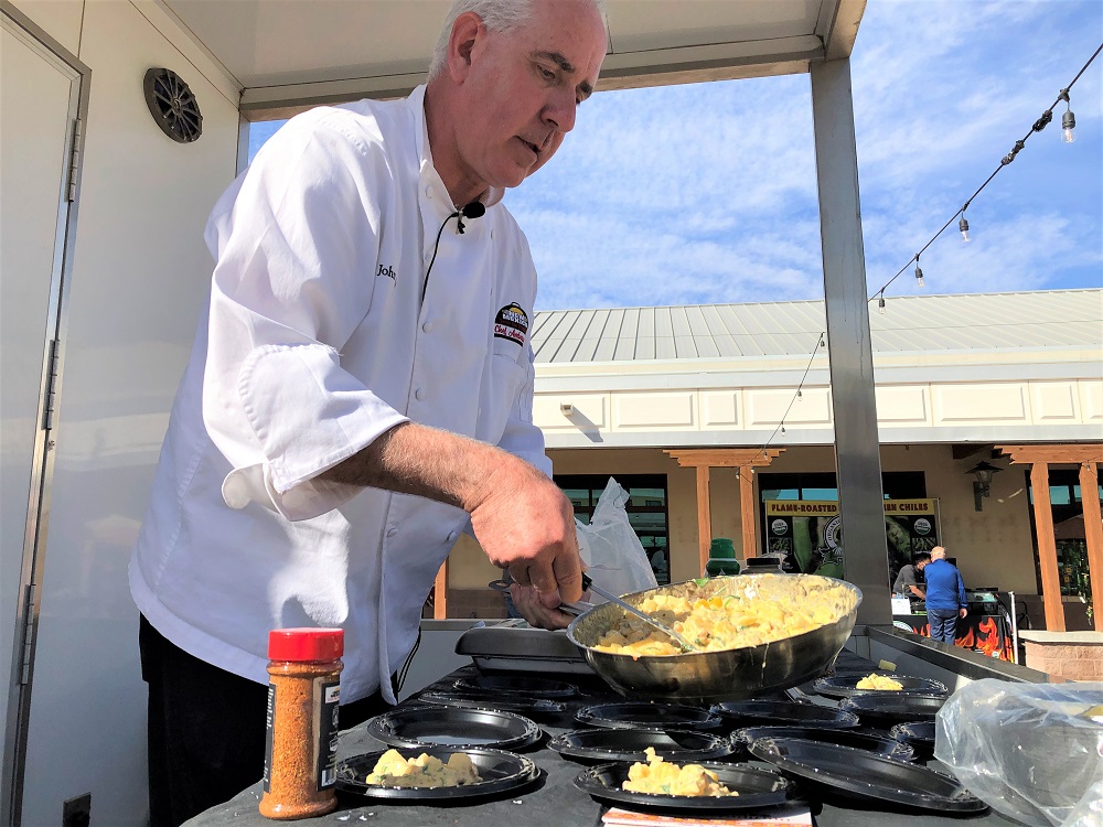 A man with a white chef’s shirt standing in a food trailer serves food from a pan onto a plate, in an outdoor atmosphere.