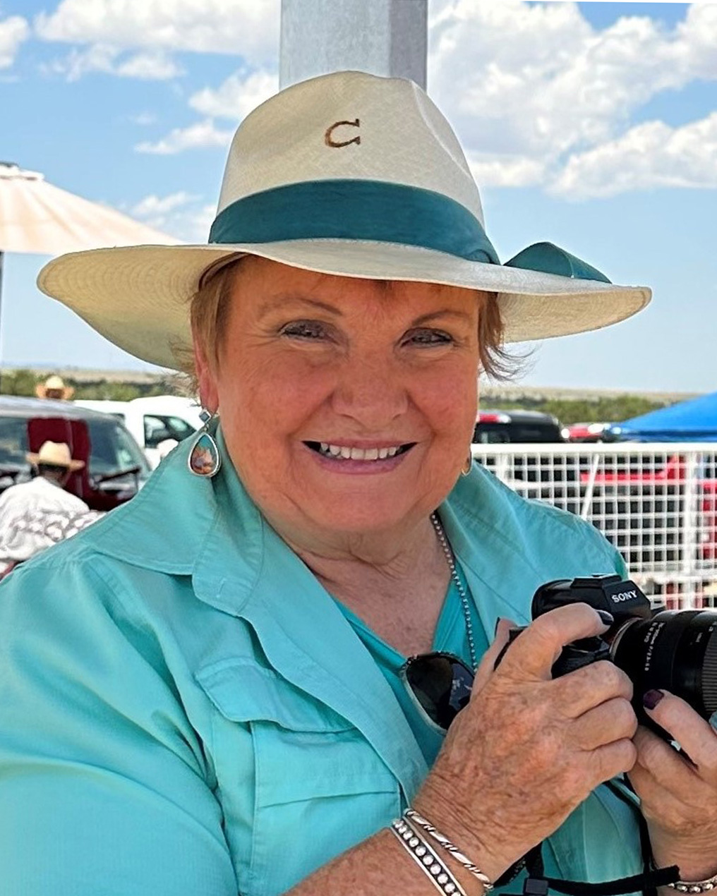 una mujer posa con un sombrero para el sol de color crema con una cinta verde oscuro y una camisa y chaqueta azul claro. Sostiene una cámara