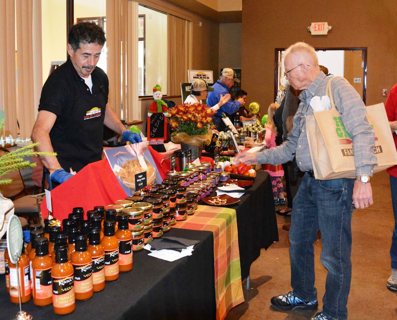 A vendor wearing gloves stands behind a table filled with bottles and jars of sauces while speaking with a customer carrying shopping bags at the HomeGrown event.