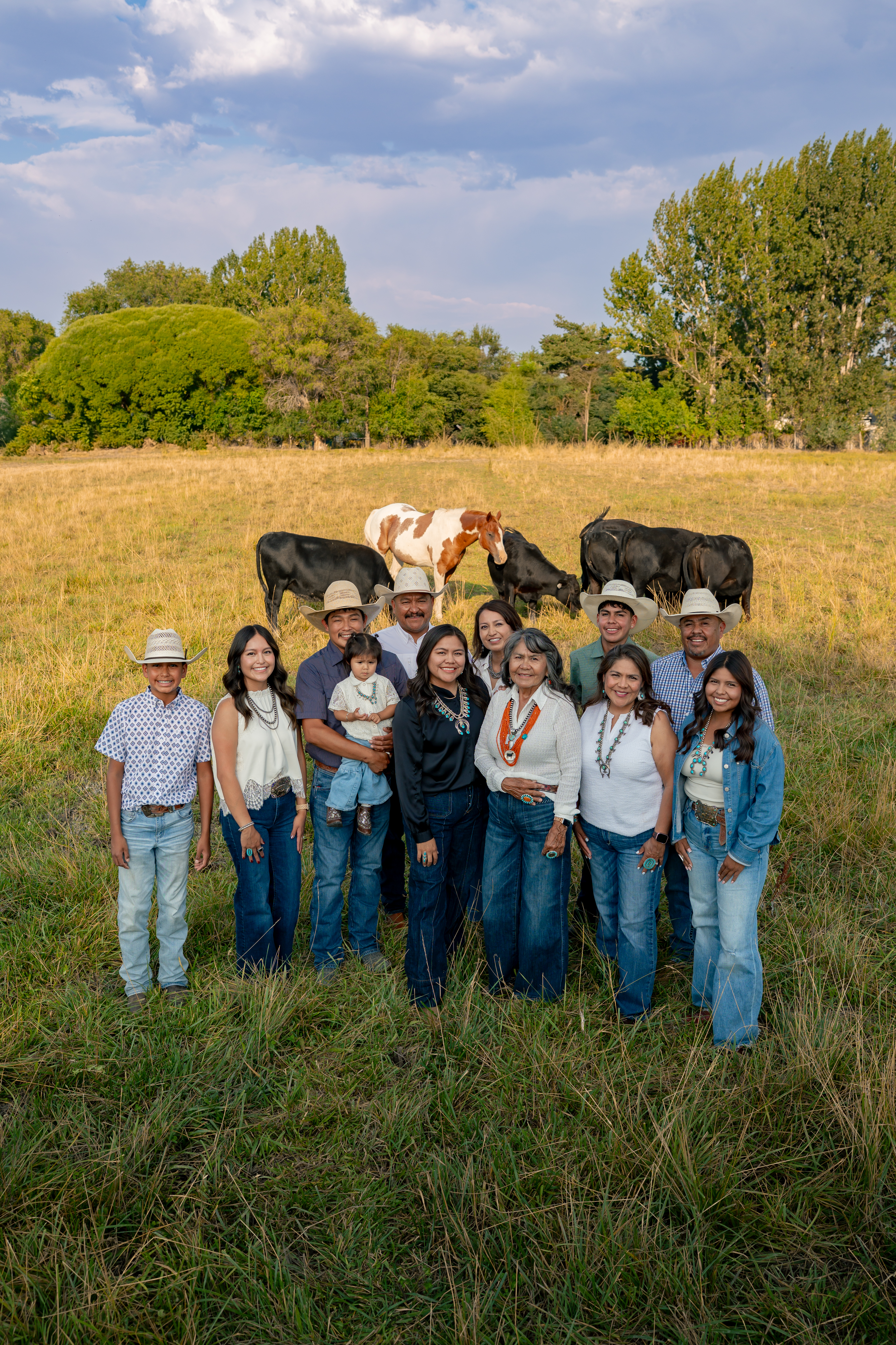 A multigenerational family stands together in a grassy pasture, with several cattle grazing behind them and trees and a partly cloudy sky in the background.
