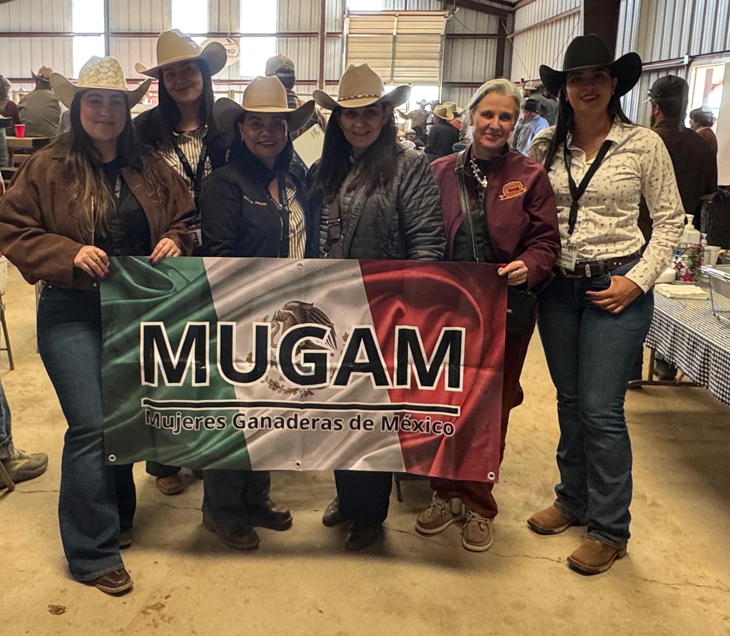 Six members of Mujeres Ganaderas de México (MUGAM), an organization of cattlewomen from Chihuahua, Mexico, stand inside a livestock sale barn holding a banner with the Mexican flag and the group’s name during a visit to a New Mexico bull sale.