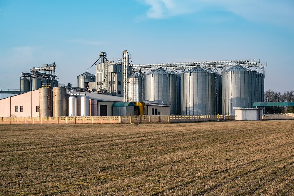 Un edificio de fabricación con silos de plata rodeado por un campo abierto con cielo azul de fondo