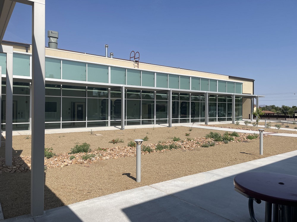 Paisaje abierto con rocas de color canela y pequeños arbustos verdes con una pared con ventanas que conduce al edificio del Laboratorio del Departamento de Agricultura de Nuevo México