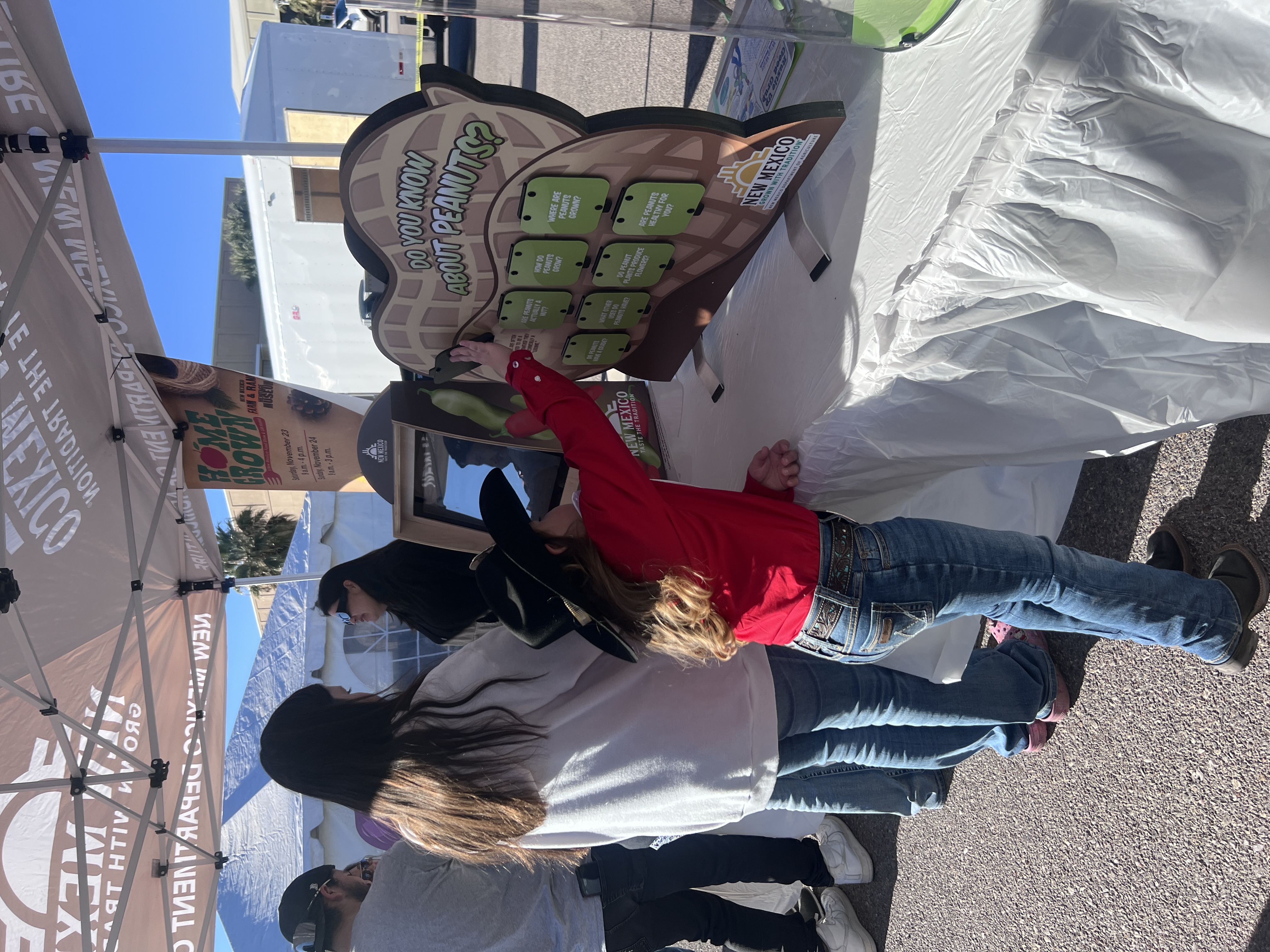 A young girl in a cowboy hat interacts with an educational display about peanuts, while an adult stands nearby under a tent at an outdoor event.