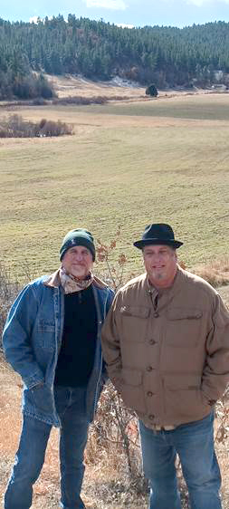 Two men wearing jackets and hats stand in front of a meadow. In the background is a forest with many pine trees.