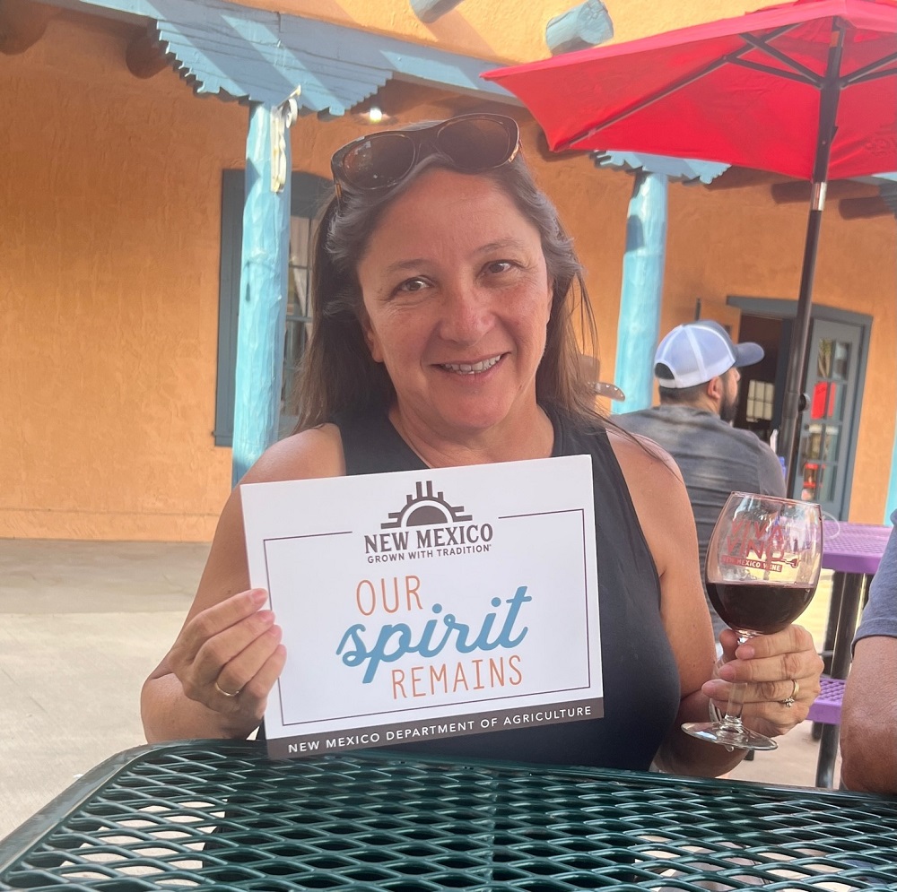 A woman holds a Viva Vino wine glass with red wine in one hand and a sign that reads, “Our Spirit Remains” in the other hand. 