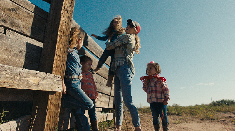 Standing next to a weathered wooden fence, a woman holds a young girl, while two young girls lean against the fence and one stands near the woman 