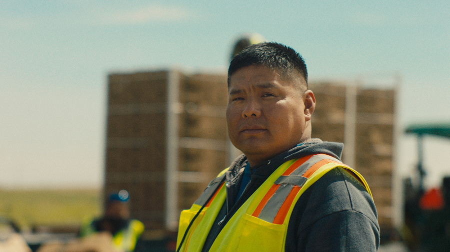 A man in a yellow safety vest looks into the camera lens, with a blurred background of a tractor, stacked boxes on a flat trailer and a field.