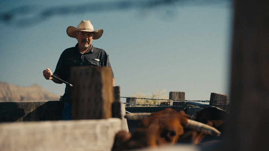 A man in a cowboy hat stands in a fenced-in area on a ranch, with a bull in the foreground.