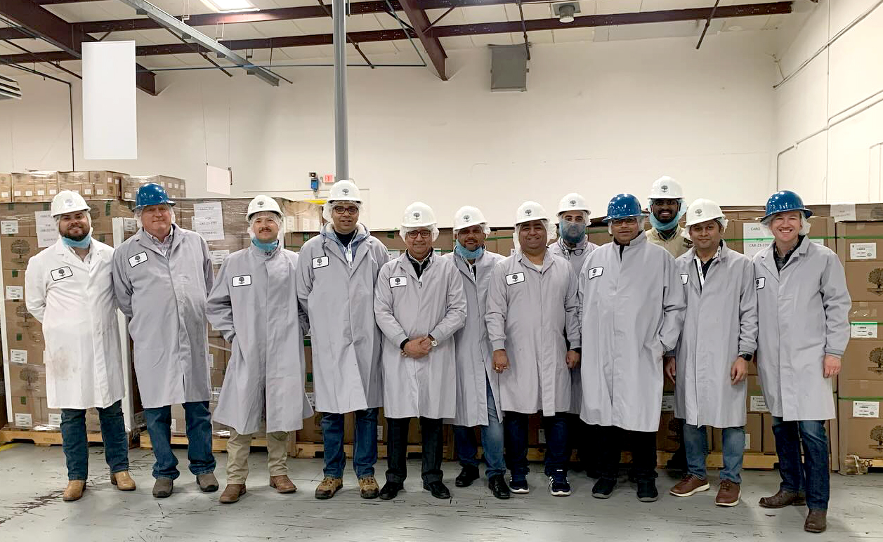 12 men in grey and white lab coats and white and blue hard hats pose for a photo inside a warehouse. Cardboard boxes are stacked in the background. 