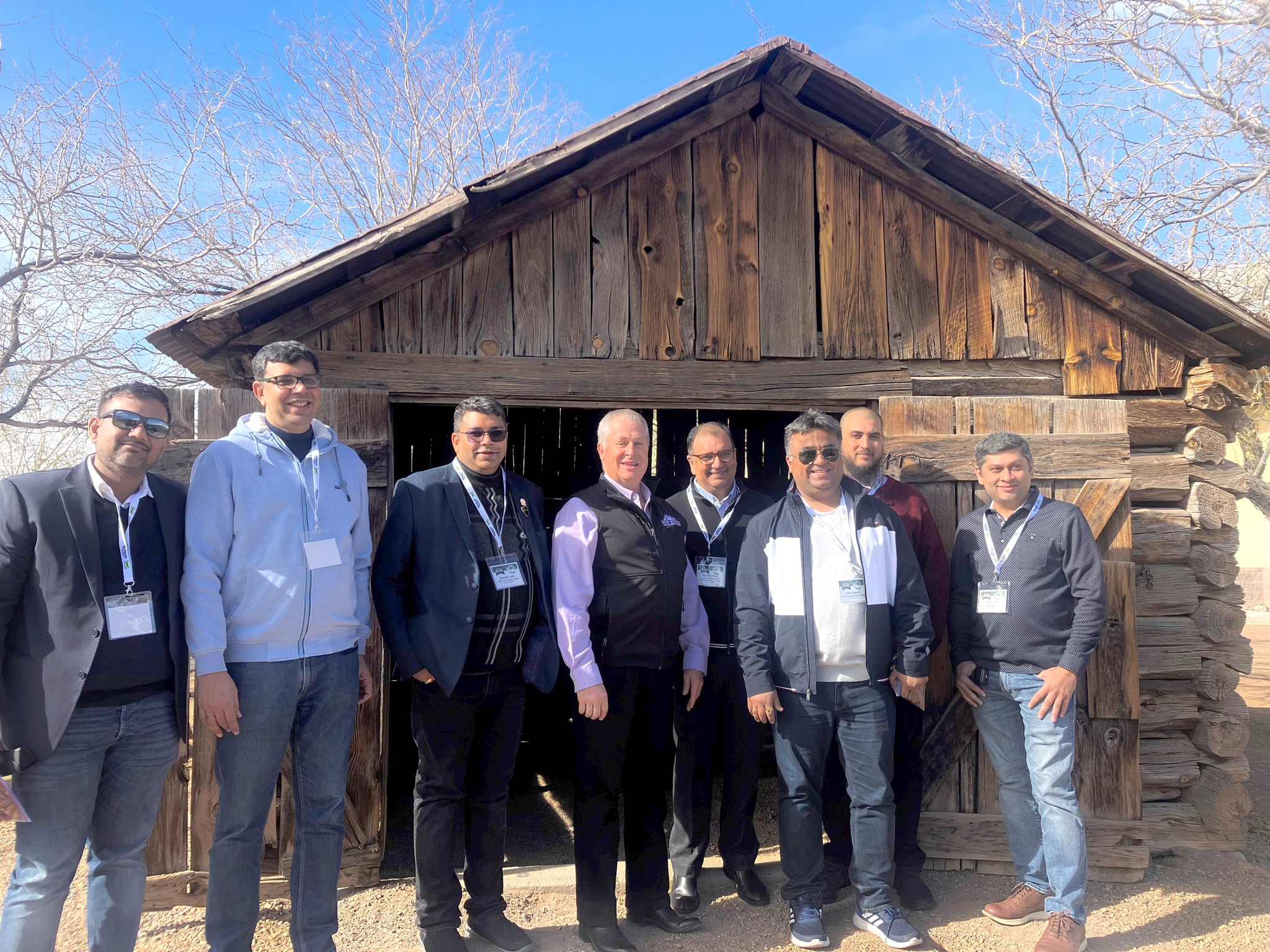 Group of eight men standing in front of an old log cabin.