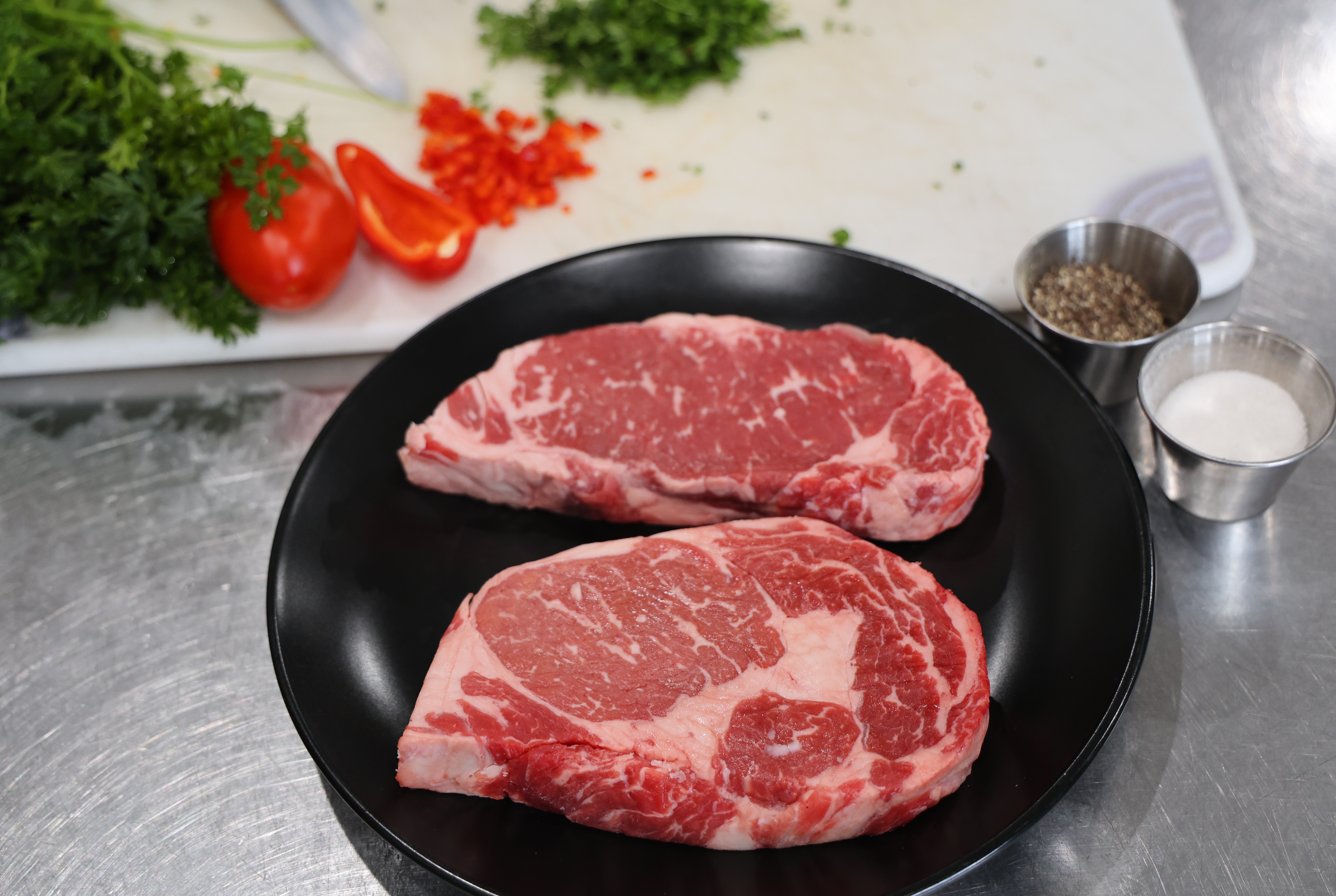 Two raw ribeye steaks on a black plate, with chopped herbs, tomatoes, peppers, salt, and pepper in the background on a prep station.