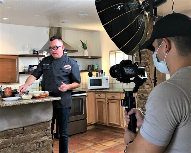 Un hombre con una camisa negra de chef en una cocina prepara un plato en el fondo, mientras que un hombre en primer plano sostiene un equipo de grabación de video.