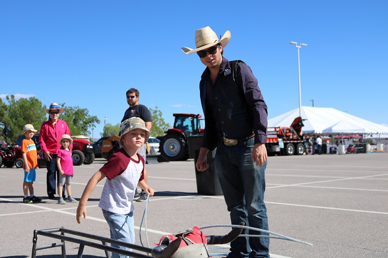 A male college student with a black shirt and cowboy hat teaches a young boy how to rope with a roping dummy. 
