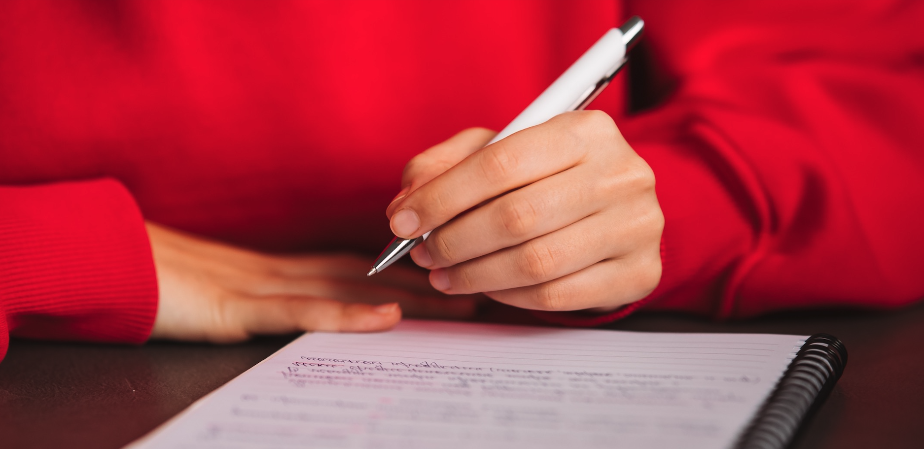 a woman wearing a red shirt writing in a notebook.