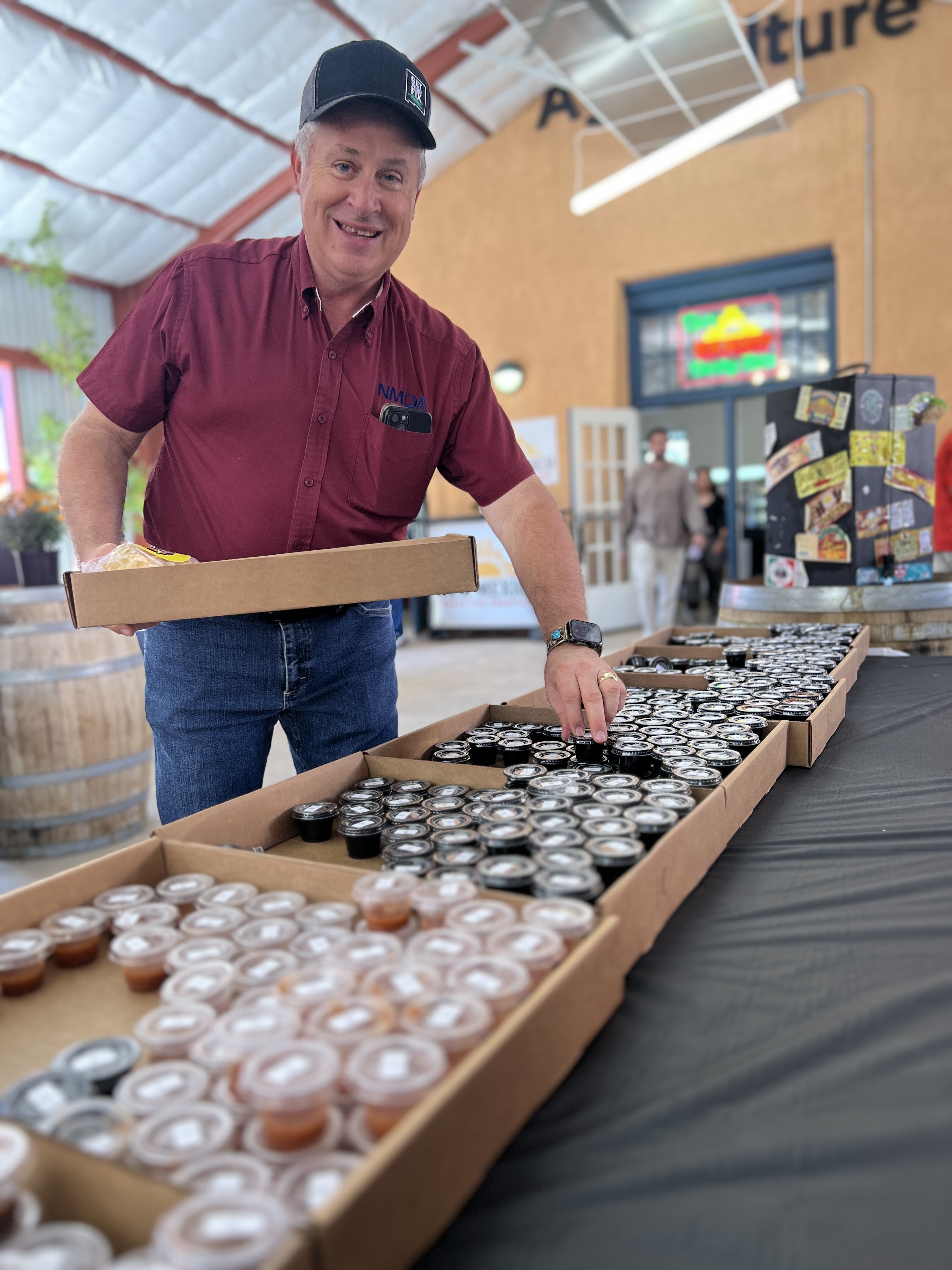 A man in a black cap, maroon button-down shirt and blue jeans picks up a tiny black single-serve cup of salsa, smiling at the camera.